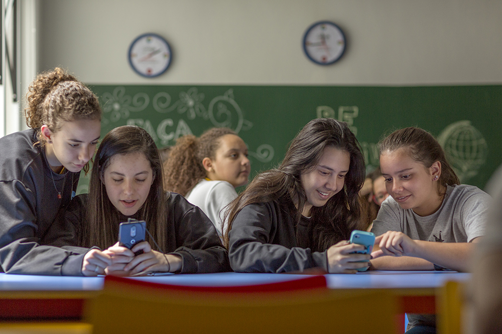 From left to right, four white teenage girls sitting behind a blue table: the first is a young redhead girl with curly hair, standing, with her head down looking at another girl's cell phone; this second girl is seated and has black straight hair, holding the cell phone in her hands; a third young girl has long, dark, straight hair and is smiling, her cell phone in her hands; a fourth girl, with dark brown hair sits next to her, interacting with the same cell phone. Behind them, in the middle, a slightly darker-skinned girl with her long curly hair up looks off to the side. In the background is a blackboard and two clocks.