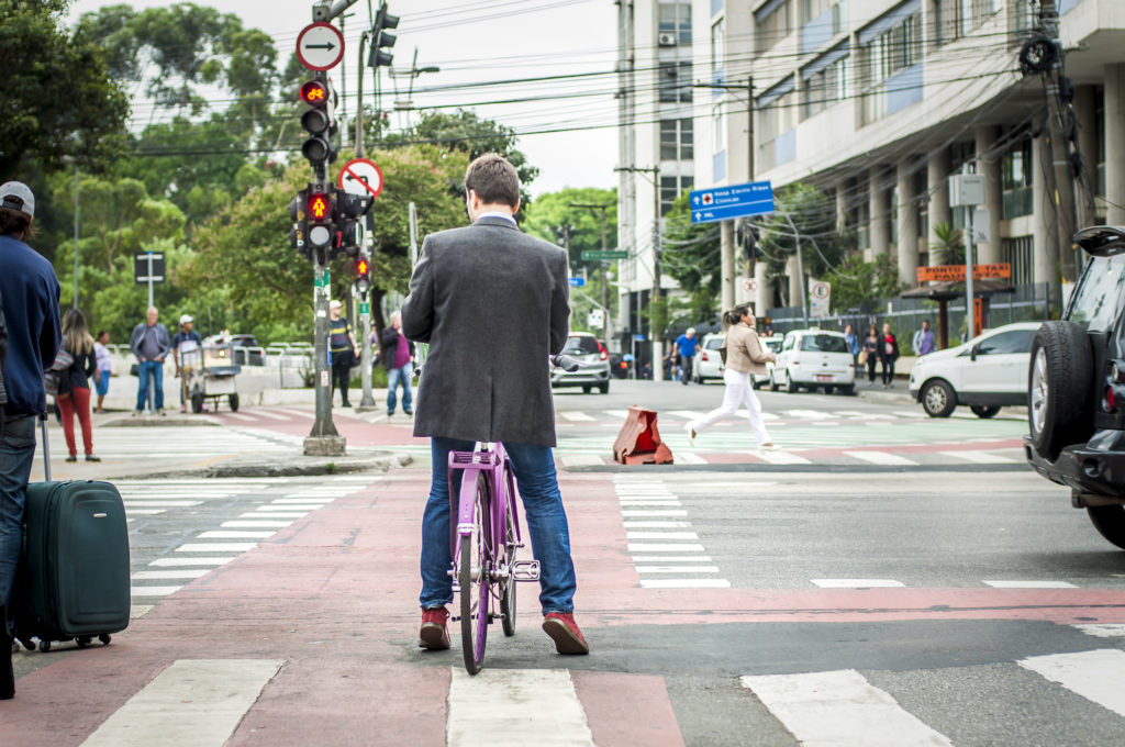 The same man featured in the cover photo, wearing jeans, a light blue shirt, a dark gray blazer and burgundy shoes is standing, on his bike waiting at the traffic light, the bike path in front of him. His back is to the camera. Busy avenues with cars and pedestrians surround him.