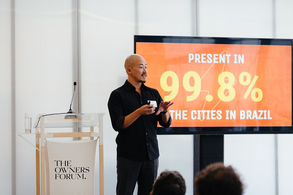 A thin, bald, Japanese man with a small beard is wearing a black shirt and black pants. He is giving a lecture. In back of him, just off to his left, a huge screen reads, "Present in 99.8 percent of the cities in Brazil". On the other side of the speaker, there is a lectern with a microphone and a sign reading,"The Owners Forum".