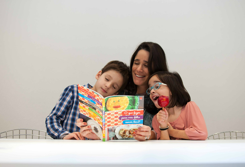 A white woman with straight hair is sitting behind a white table (we only see the top of the table). She is holding open the book "How Can I Get My Child to Eat Well?" She is smiling and looking startled. There are two children, one on either side of her. A white boy, around the age of 7, rests his head on the woman's shoulder, his eyes closed. The other child, a girl with black straight and short hair, holds a candy apple and wears blue-rimmed glasses. They are all looking at the book together. The little girl’s eyes and mouth are wide open as she notices, with delight, something on the page.