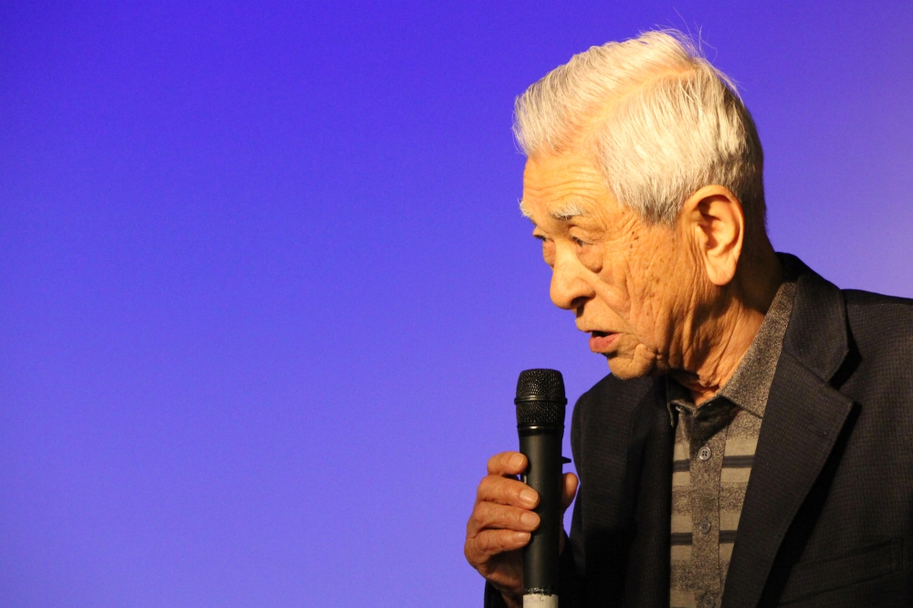 Against a blue background, a Japanese man, about 80 years of age, holds a microphone. He is speaking, presumably to an audience. He is shown in profile from the chest up.