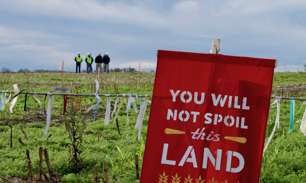 Uma bandeira vermelha com a inscrição, em inglês, "Vocês não vão envenenar essa terra", está fincada em um campo verde à frente de uma cerca. Atrás, um grupo de quatro homens, usando roupas e capacete de segurança, está conversando. 