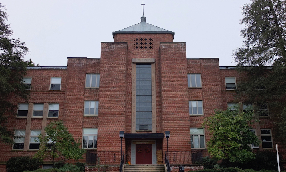 A three-story brick building, with a cross on the roof at its peak. At the entrance, a stairway of about ten steps offers access to the main door (the only one that appears in the image). The building also has a set of glazed windows and is surrounded by trees and shrubbery.
