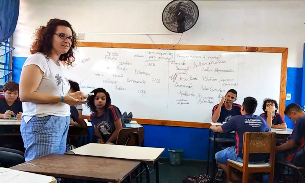 Uma mulher jovem, branca de cabelos cacheados e óculos está em uma sala de aula. Em pé ela parece explicar algum assunto aos alunos, que estão sentados olhando para ela. Ao fundo, um quadro branco com várias anotações.
