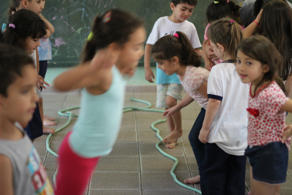 Um grupo de sete crianças, com mais ou menos 8 anos, formam duas fileiras, frente a frente. Há uma corda no chão que as separa. E uma delas, que aparece desfocada na imagem, está pulando por cima dessa corda.