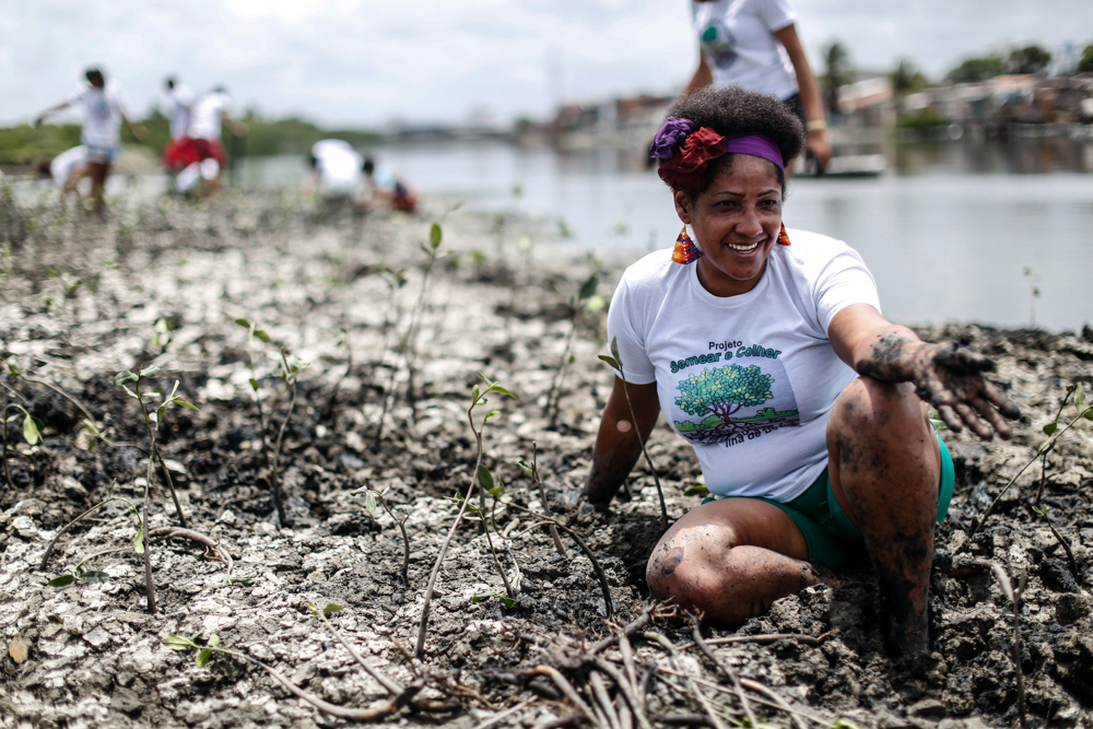 A black woman with short hair trimmed with a strip of lilac and red flowers wears colorful earrings and a white T-shirt bearing the slogan "Plant and Harvest". She is sitting and smiling, her feet sunk into the mud. Behind her, the dark river and some people out of focus.