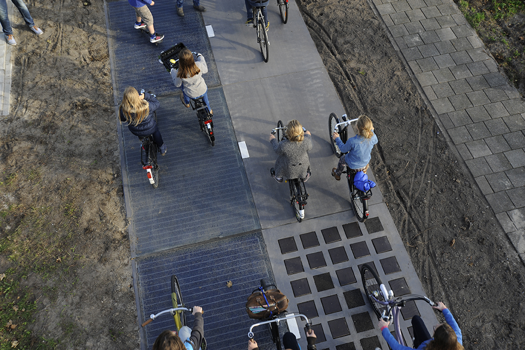An aerial view of a group of about eight people riding bikes on asphalt. Part of the sidewalk is covered by a set of black squares.