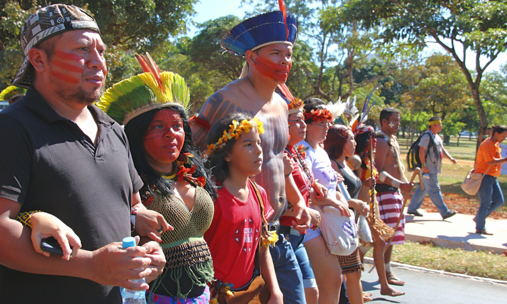 A foto mostra Sonia Guajajara com o rosto pintado e usando cocar, com outros 7 indígenas, entre adultos e crianças, também usando cocar e com o rosto pintado. Eles estão com os braços entrelaçados em uma fileira horizontal e caminhando em uma estrada asfaltada. 