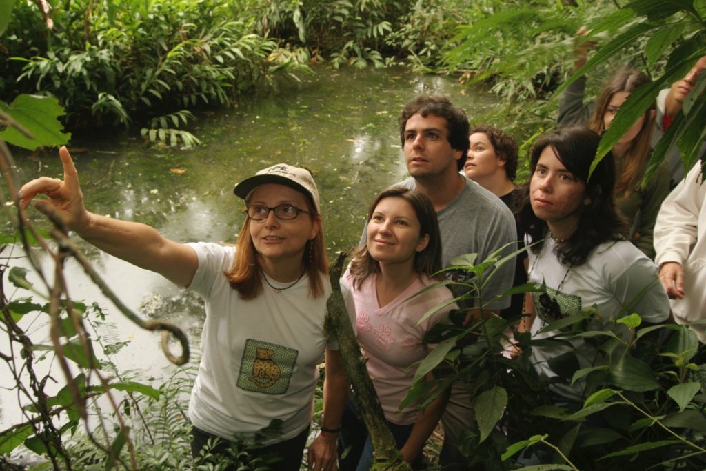 The photo shows a woman with brown hair down to her shoulders, wearing a cap, glasses, and a white shirt. She is pointing to a tree on a trail, next to a creek, followed by a few people who look in the direction she is pointing.