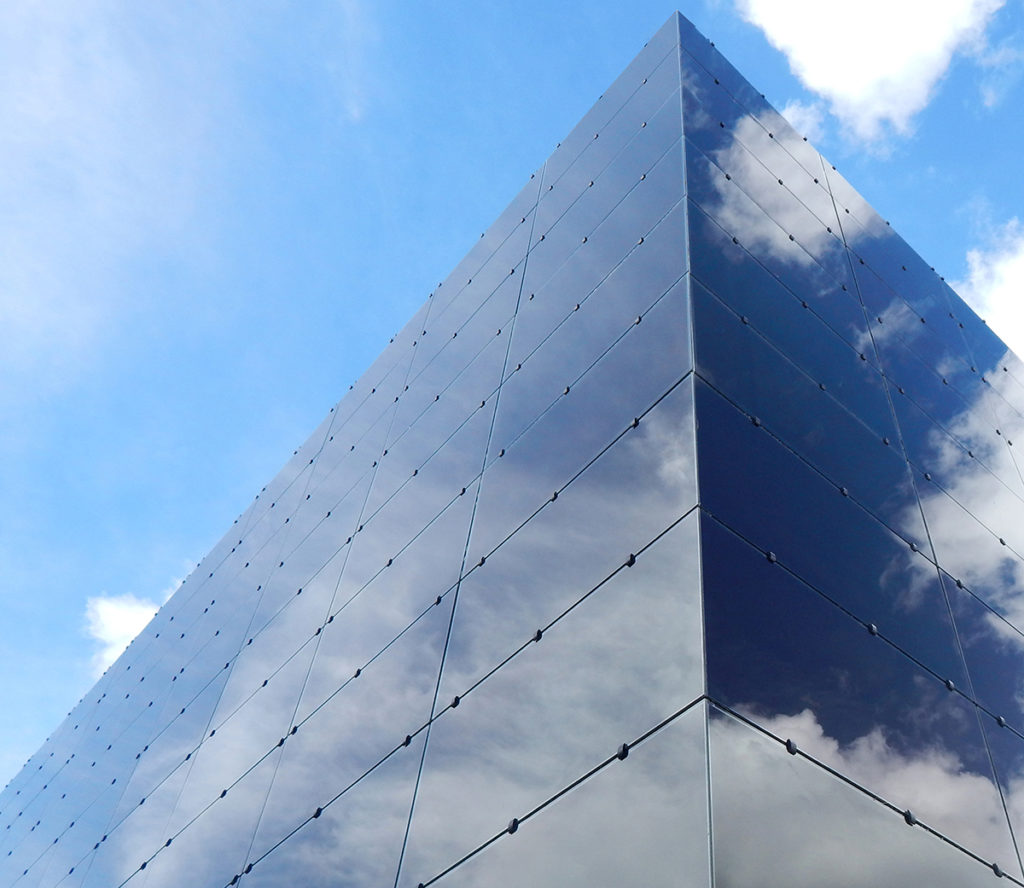 Imagem em plano fechado da diagonal de um edifício feito todo de blocos retangulares de um vidro com um tom escuro de azul. Por este vidro vemos o reflexo do céu azul com nuvens. A imagem mostra até o topo do prédio, o céu azul e algumas nuvens.