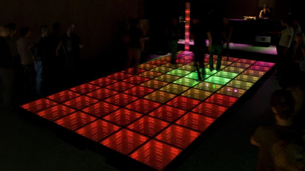 There is a rectangular dance floor formed by several colored squares, which light up in red, green and pink. On top of it, people are dancing, their images blurry.