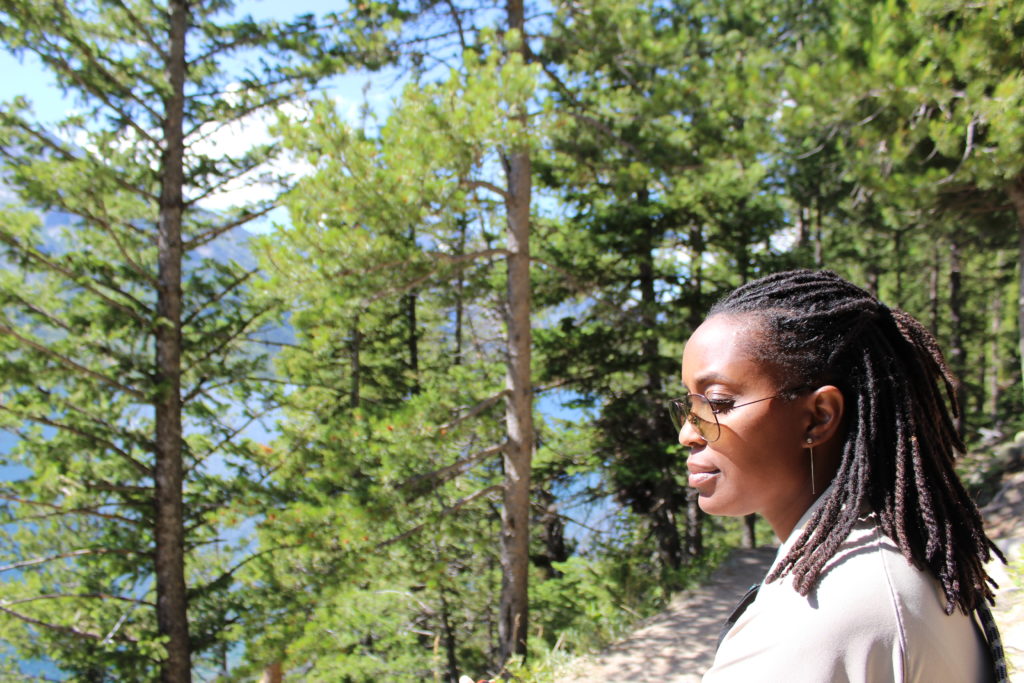 In the middle of a pine forest, a black woman in profile, wearing a cream-colored shirt and glasses, her hair in braids, looks ahead. 