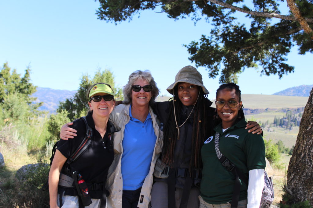 Four women are embracing, surrounded by trees, open space and sky. A white woman on the far left wears a green visor, sunglasses and a fanny pack. To the right of her is a second white woman, this one gray-haired, wearing a blue shirt and black sunglasses. To the right of her is a black woman with long braids wearing a brown hat. To the right of her is a second black woman, her braided hair tied back, wearing big glasses.