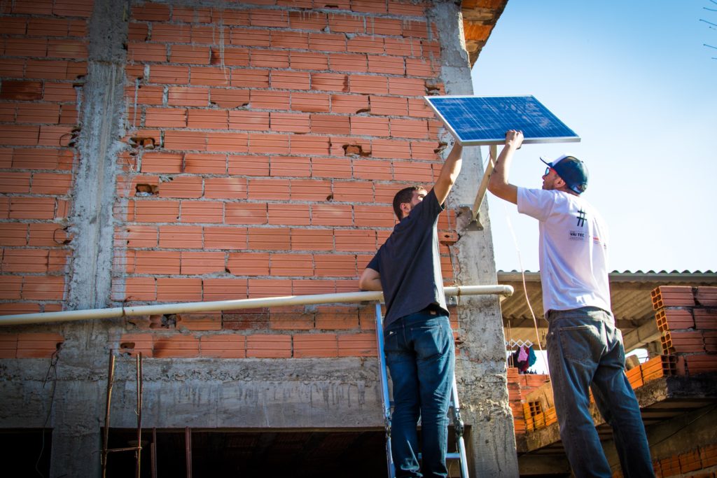 A imagem mostra em plano aberto um grande muro de tijolo à vista, onde dois homens magros, vestindo jeans e camiseta estão instalando uma placa fotovoltaica pequena (menos de 1 metro quadrado). Eles seguram a haste da placa e estão de costas para a imagem.