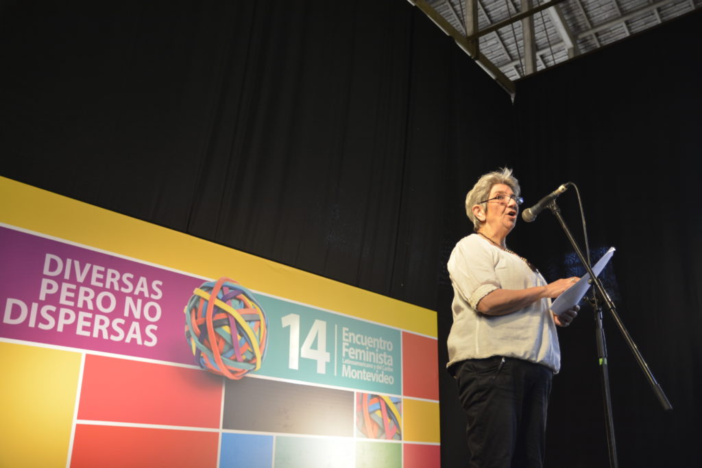 A short, gray-haired white woman wearing glasses, a white shirt and black pants. She is speaking into the microphone, standing on a stage. Behind her is a large banner that reads, in Spanish, "14th Montevideo Feminist Meeting - Diverse but not Divided."