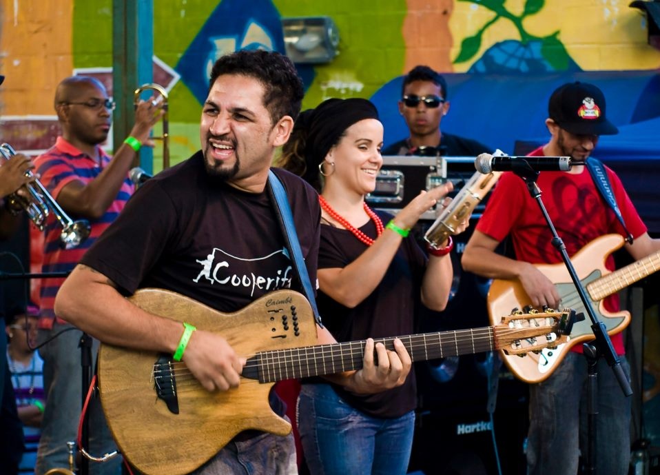 On the left is a dark-haired man with a goatee. He is holding a guitar and looking to the left. Next to him is a white woman with brown hair wearing jeans and a black T-shirt. She is playing the tambourine and smiling. Next to her is a thin man, wearing jeans, a red shirt and a cap. He is looking down and playing the bass. Two other musicians, behind them, play some sort of percussion instruments. 