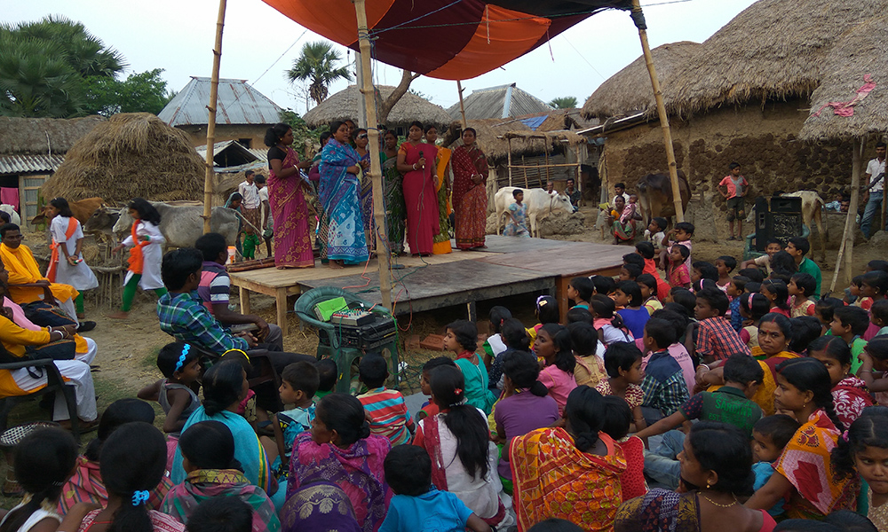 There are seven Indian women wearing long and colorful sarees (typical dresses of their culture) standing on a makeshift wooden podium covered by a canvas roof. In the foreground, women and children, their backs to the camera, face the podium, sitting and watching. In the background are several houses and huts made of clay, some with thatched roofs. 