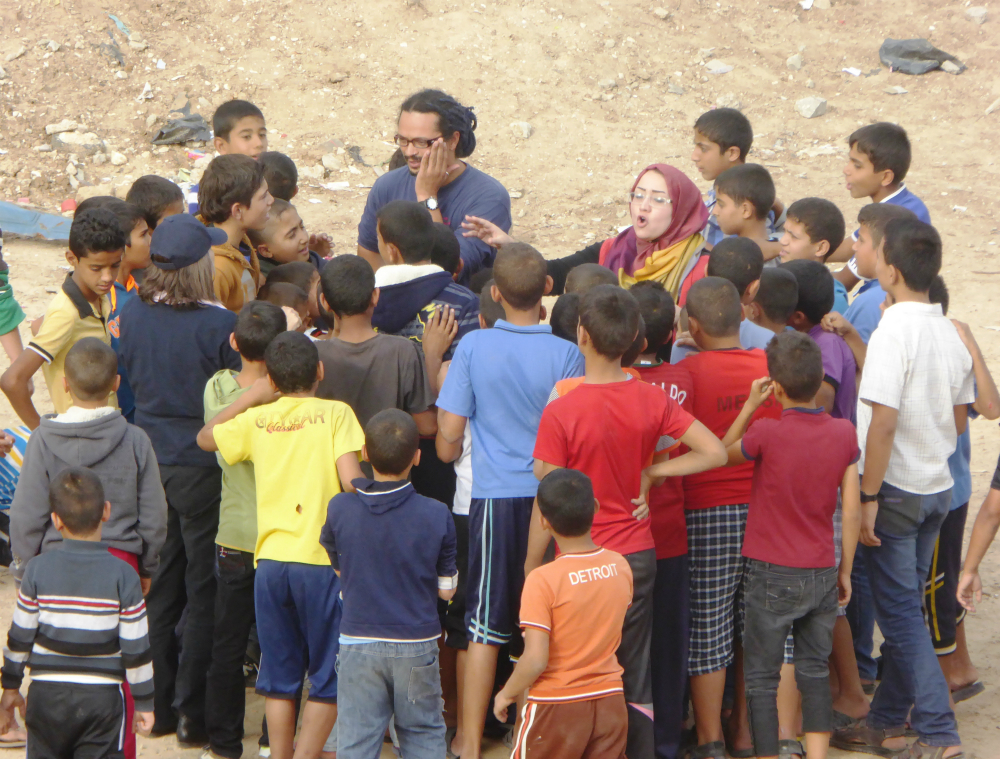 The man described previously is surrounded by children. Next to him is a white woman wearing a scarf on her head, gesturing and speaking, perhaps singing. In the background, parched-looking dirt and rocks.