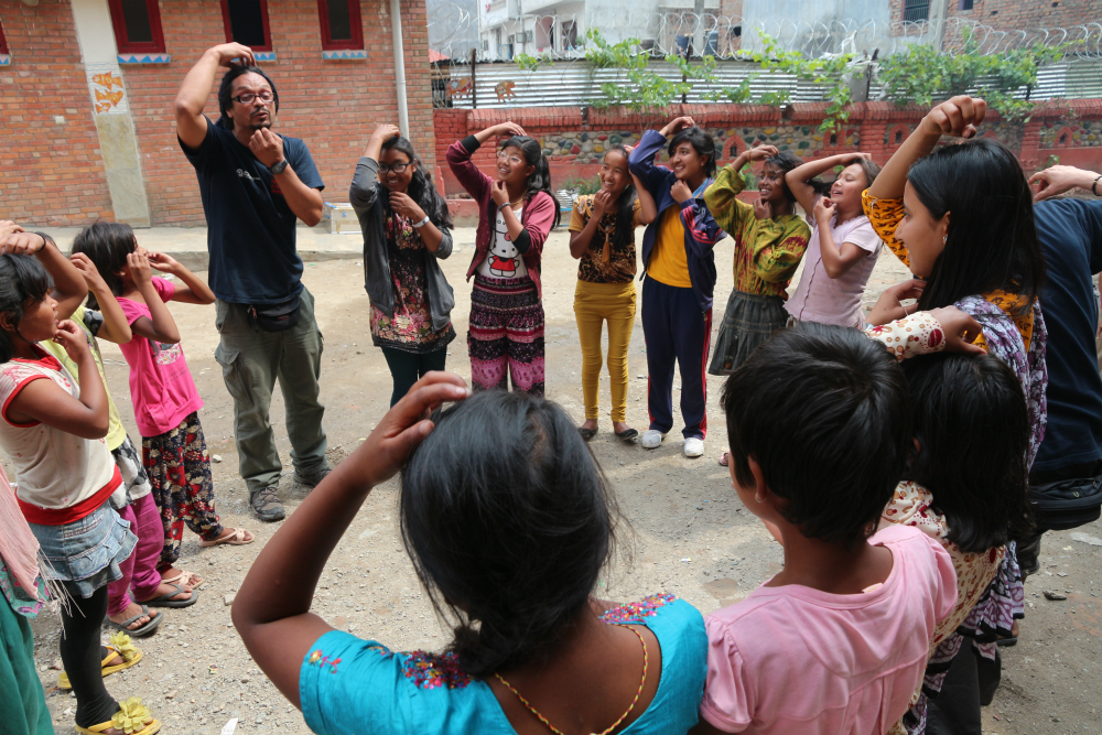 The same man described in the previous photo stands in a circle with several brown-skinned, black-haired children. They seem to be playing a mimicking game: the kids are looking at and imitating the man, whose right hand scratches his forehead while his left hand scratches his chin.