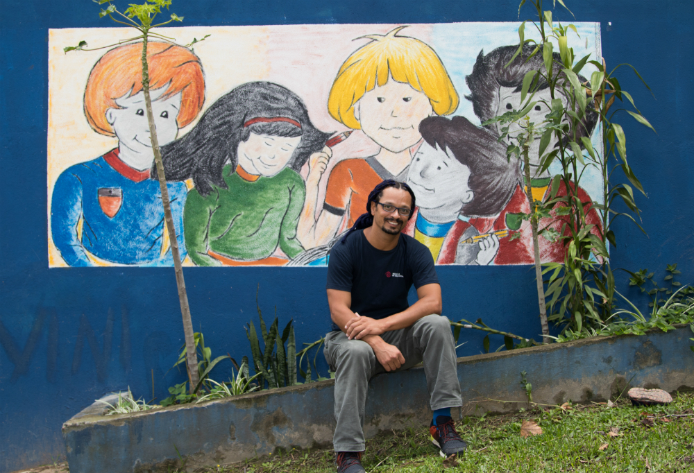 The man described previously is sitting on a short wall, his feet resting on a green lawn. In the background, a mural depicts children with orange, black and yellow hair. 