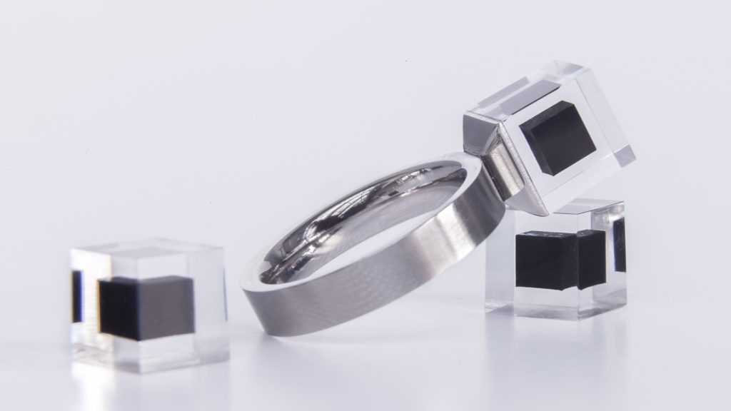 A metal ring, and attached to it, a square transparent "stone,” which looks like acrylic or clear glass, with another small black square inside of the clear one. Two other identical stones lie next to it. 