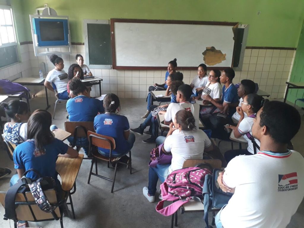 A foto mostra um grupo de adolescentes conversando em uma sala de aula. Os jovens estão sentados em carteiras e cadeiras, e ao fundo há uma televisão, uma lousa e, sobre ela, um quadro branco.