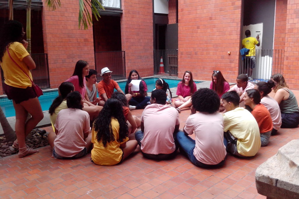 A group of teenagers is sitting in a circle in a courtyard with an brick-orange tiled floor. In the background is a pool, and walls covered in tiles the same color as the courtyard floor.