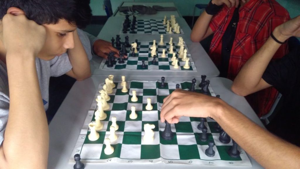 La foto muestra tres tableros de ajedrez, uno junto al otro, en una mesa larga, en torno a la que están sentados algunos jóvenes que juegan.