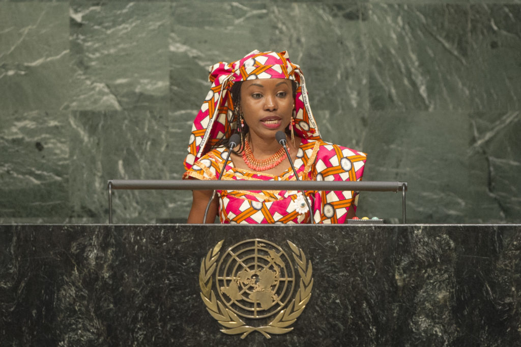  A black woman, wearing a dress and matching headscarf in a red, white and orange pattern, is speaking from a lectern, upon which is the emblem of the United Nations.