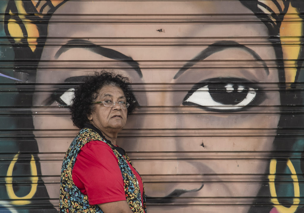 A black woman with curly hair, turned sideways. She is wearing glasses, a red shirt and a colorful, flowered vest. She is looking at the camera without smiling. Behind her, a mural depicting the face of a black woman with big eyes.