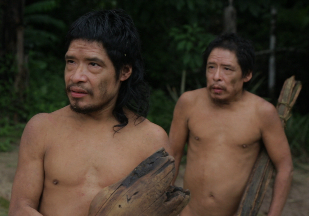 Two short, brown-skinned, shirtless men (shown from the waist up) are looking to their right. The first man, in front of his companion, has straight, black, shoulder-length hair and a slight beard and mustache. The second man looks a little like the first one but seems to be older and with shorter hair. Both are carrying firewood.