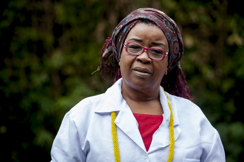 A black woman, shown from the waist up, with long braided hair, is wearing a printed turban, red rimmed glasses, a yellow necklace and white lab coat. She is looking at the camera. The background, of leafy greenery, is out of focus.