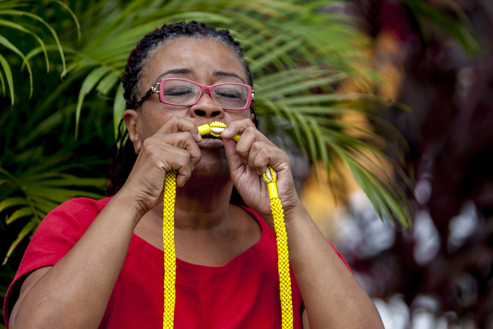 A black woman with long braided hair, tinted red, wearing red-rimmed glasses. Her eyes are closed, and she holds a necklace made of yellow beads, kissing it.