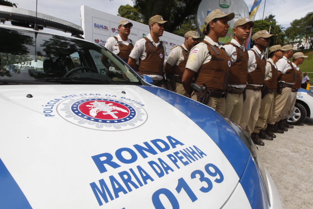 In the foreground, the hood of a police car, bearing the police force’s coat of arms. On the hood are the words, "Ronda Maria da Penha 0139." In the background, a group of more than 10 police officers is lined up in 3 horizontal rows, standing at attention. They are all wearing dark brown vests, with beige pants and matching beige caps. 
