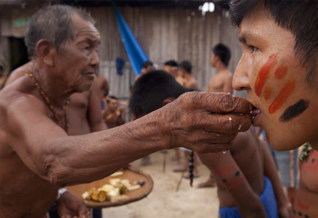In the left corner of the image, an elderly indigenous man, shirtless and wearing a colorful bead necklace, looks to the right side of the image as he uses his right hand to put food in the mouth of another indigenous man. This man is younger, with his face painted in red and black stripes. In the back are other indigenous men, out of focus.