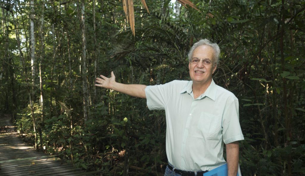 The same white-haired 66-year-old man from the previous photo is wearing glasses and a light blue shirt. His right arm is outstretched, his hand pointing to the forest behind him. In his left hand, he holds a blue folder.
