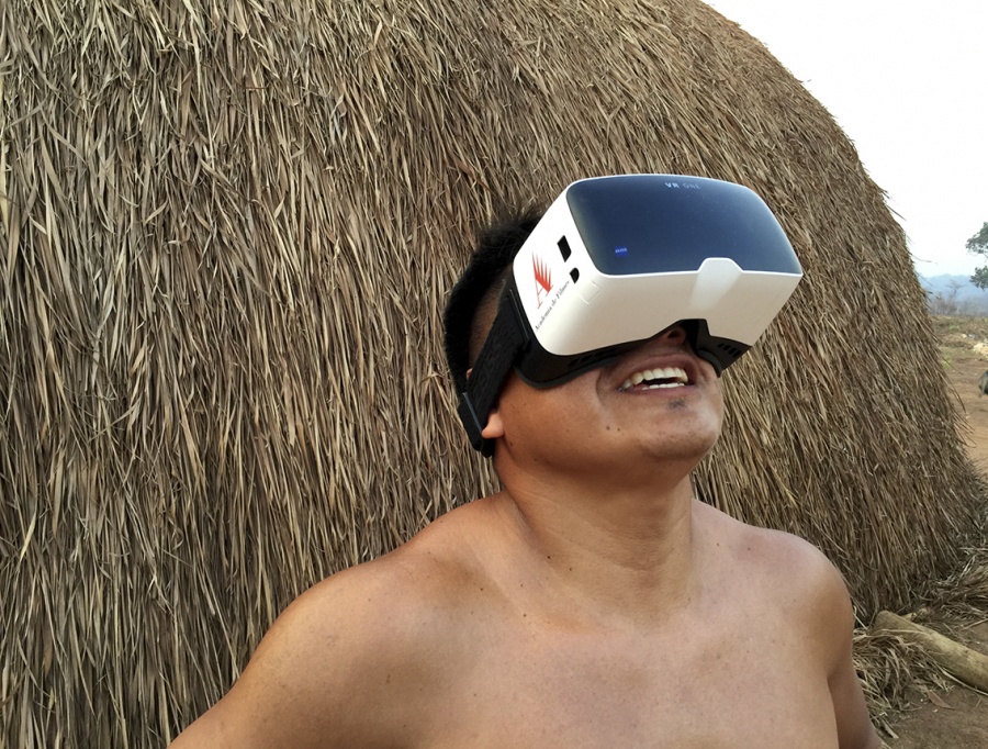 A brown man with short, dark hair is wearing, tied around his head, a virtual reality headset, which has a white plastic base, with a large dark lens covering his eyes and nose. Head lifted slightly, he smiles. Behind him is a straw hut, only partly shown.