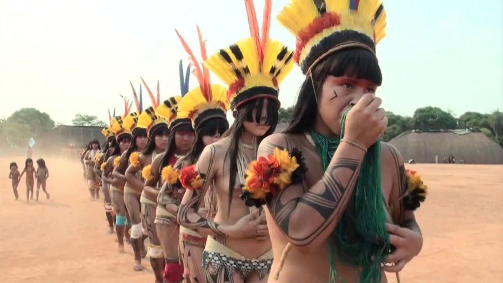 More than 10 indigenous women form a line, wearing headdresses made of red and yellow feathers, with small feathers tied around their painted arms, and wearing necklaces that cover their breasts. They are walking and seem to be dancing in a ritual.
