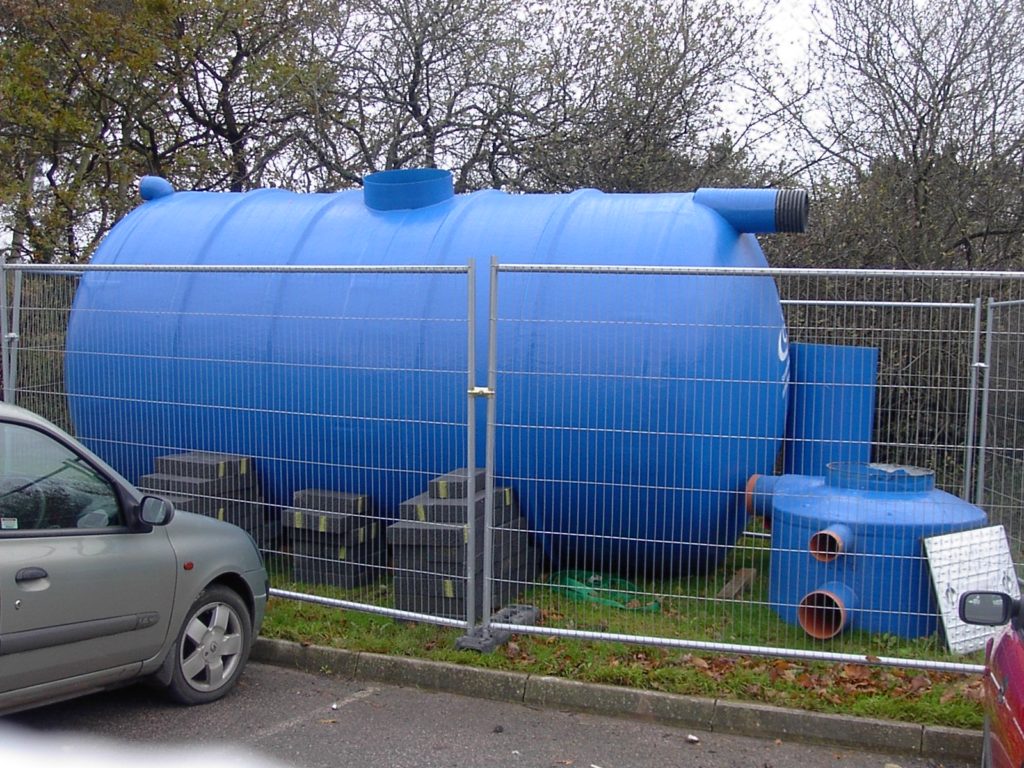 On a lawn, behind a fence, is a blue, plastic, cylindrical tank with some cinderblocks stacked against its side to stabilize it. Next to the tank are other plastic parts apparently not in use at present. In the foreground, in the left corner of the photo, is a parked car. The tank is much larger than the car. 
