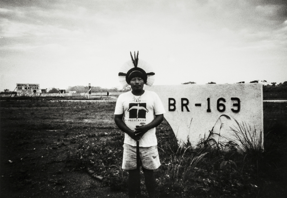 Black and white photo of an indigenous man, wearing a feather headdress, jean shorts and a white t-shirt, posing in front of a sign that reads "BR-163."
