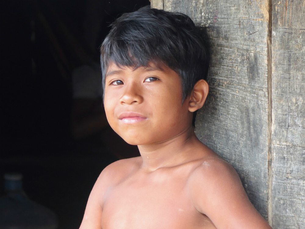 A dark skinned indigenous boy, with short black hair and brown eyes, shirtless, is resting against a wooden wall. He is looking straight at the camera. 