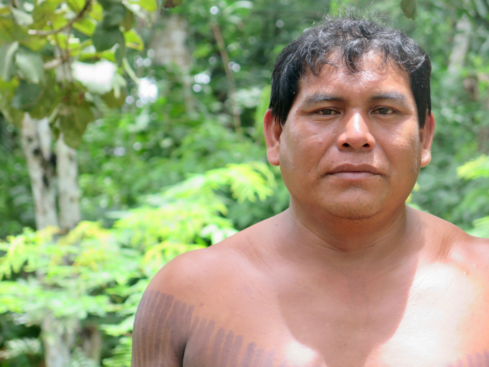 A man, seemingly between 30 and 40 years old, with dark skin, short hair, shirtless and with part of his chest painted with black stripes. He looks at the camera without smiling. Behind him are some trees out of focus.