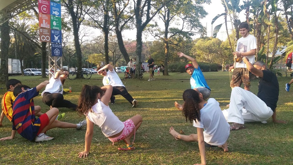 Children and adults are in a circle in a grassy field, doing capoeira movements, with their left hand on the ground, the left leg stretched forward, the right leg flexed back in a squatting motion, and their right arm over their heads. In the back is a scaffolding with colorful signs, each with a different vibrant background and the title of one of the SDGs (Sustainable Development Goals) established by the United Nations. Further back are some trees, a few bicycles, and some people walking around.