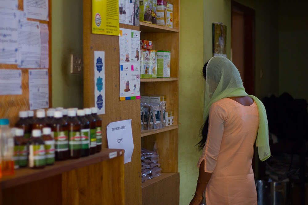 A woman with her back to the camera looks at a variety of packaged products, displayed on a light colored wooden shelf, in the center of the photograph, facing diagonally towards the camera's right corner. The woman is wearing a pink dress with sleeves down to her elbows and a see-through green veil over her long, straight dark hair.