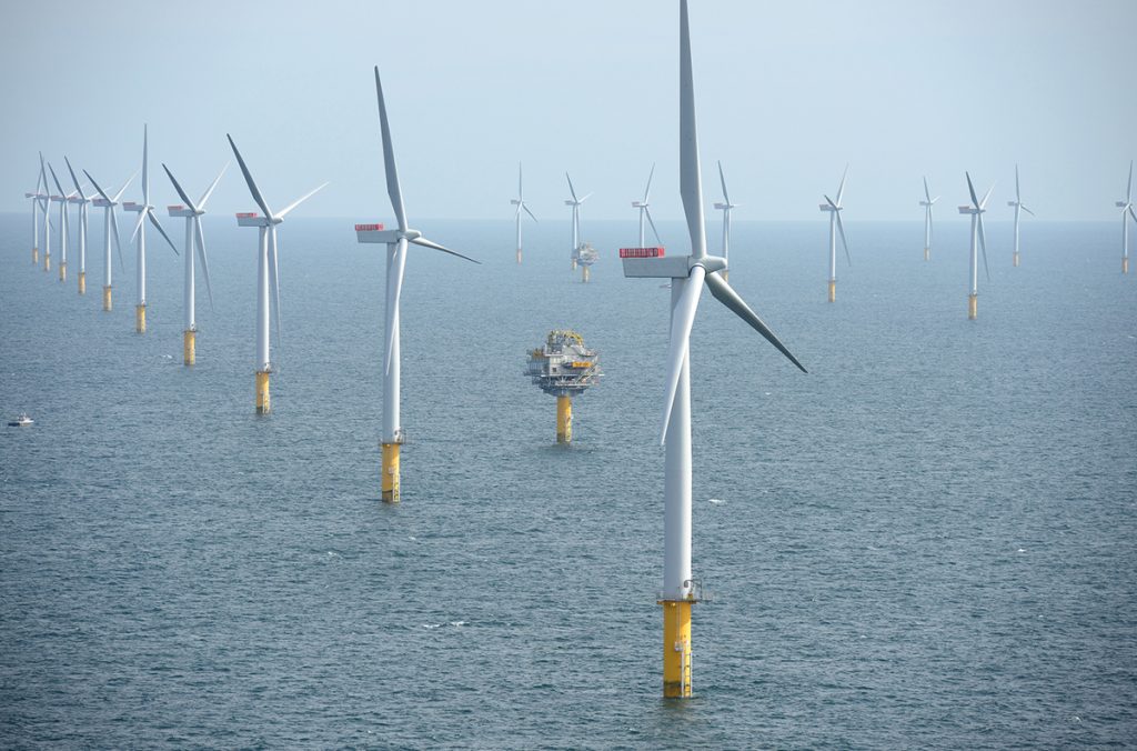 Tall, white wind turbines, installed in rows in the middle of the sea. In the background, a blue-gray sky.