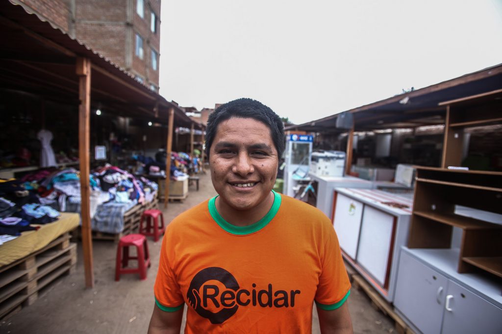 A Peruvian man, with brown skin, smiles at the camera. He has short, dark straight hair and is wearing an orange t-shirt with a green collar, with the "Recidar" logo printed in black on the chest. Around him and in the back are stands with furniture and items of clothing arranged in them, and a gray sky.