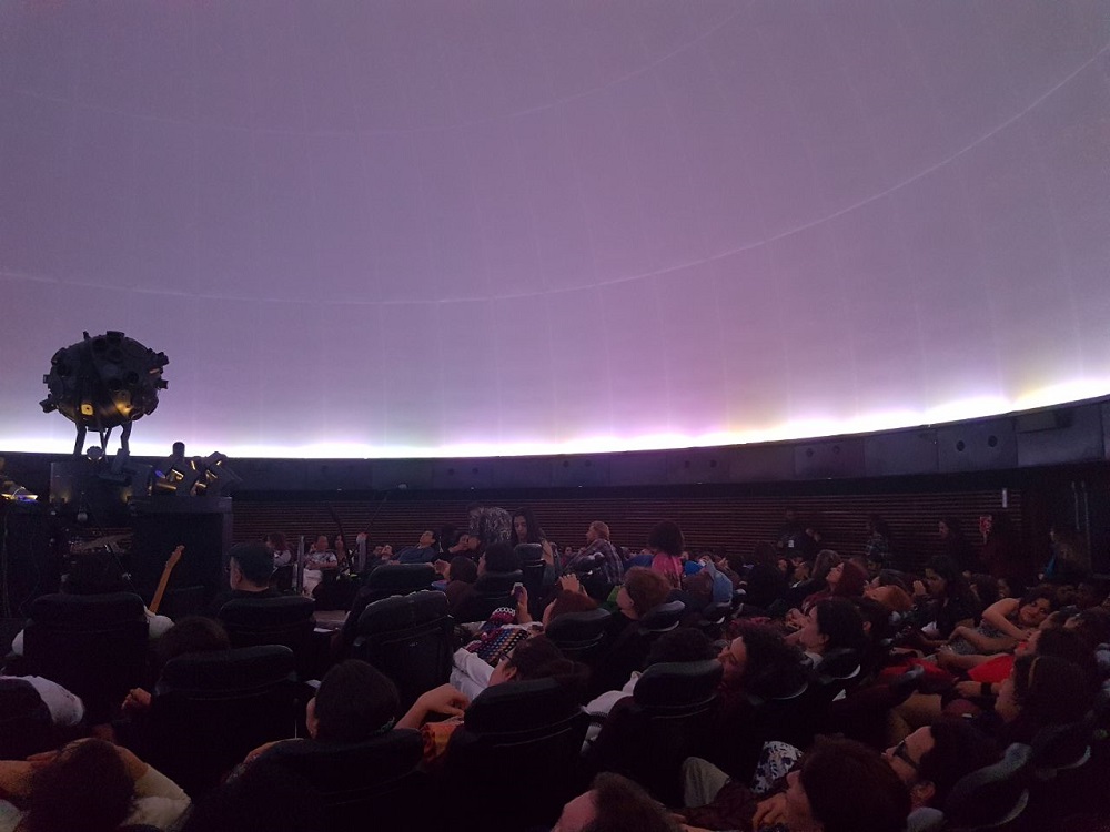 People in dark reclining seats are watching an exhibition on a semi-spherical screen attached to the ceiling. On the screen, a blueish background is surrounded by a fine bright line. In the mid-left part of the photo is a spherical image projector.