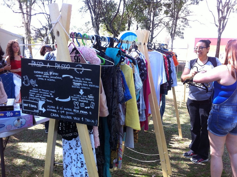 A wooden clothing rack displays various pieces of clothing, with a dark chalkboard on which directions for acquiring the items is written. The text is small, illegible in the photo. In the back are some women standing, some trees, and a lawn.