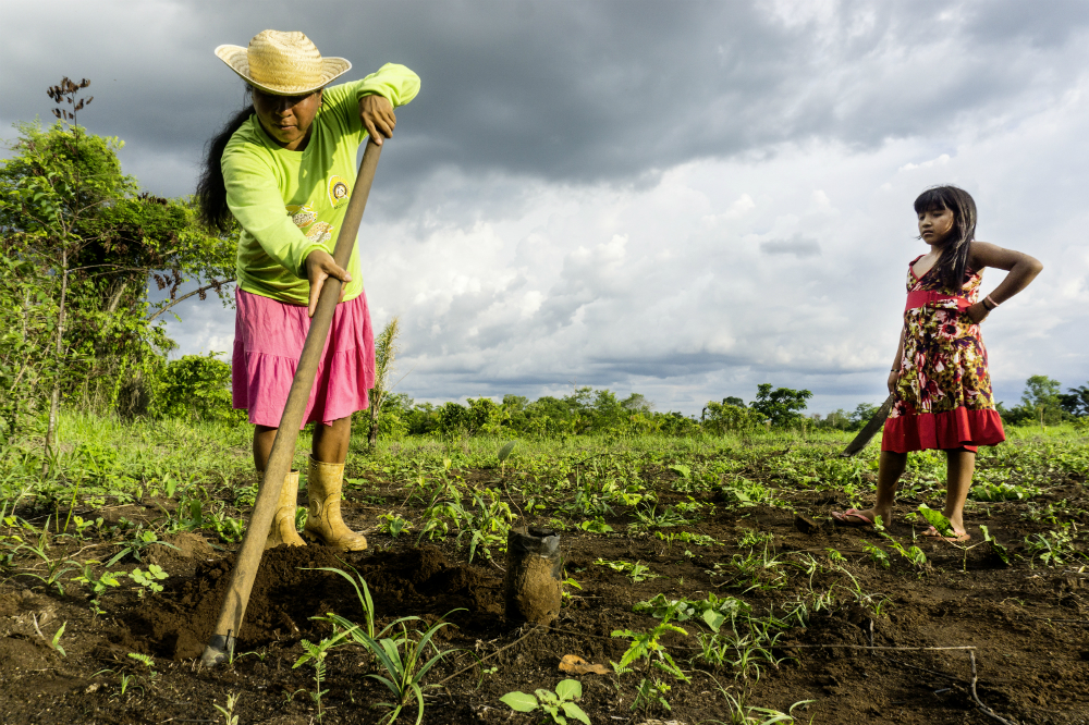 Near the left edge of the photo, an indigenous woman facing the camera is holding a large hoe that is digging into the earth in front of her. She has long, straight dark hair in a ponytail, and wears yellow rubber boots, a pink skirt down to her shins, a lime green long-sleeved shirt and a straw hat. In the right side of the photo, an indigenous girl watches the woman work, her left hand resting on her hip. She has long, straight dark hair with bangs that cover her forehead. She is wearing a flower print dress that is yellow and red, with red trim and a red band at the hip, and pink flip-flops. The woman and girl are standing on soft, dark earth and thin green grass. In the back is a cloudy sky.