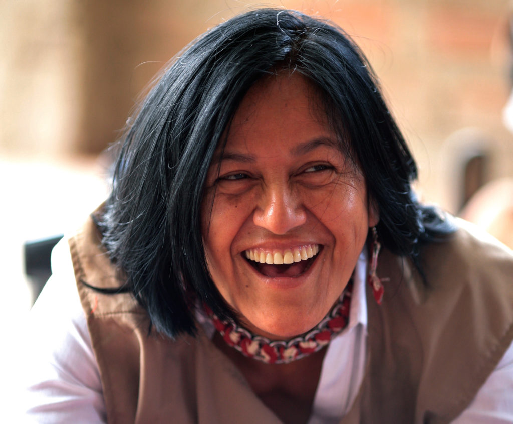 The photo shows the face of a woman with bronze skin, straight black hair down to her shoulders and dark brown eyes. She is wearing a light brown vest over a white shirt and has a red necklace. The woman is facing the camera but is looking off to the right of the photo. She has a wide open smile, exposing her upper teeth.